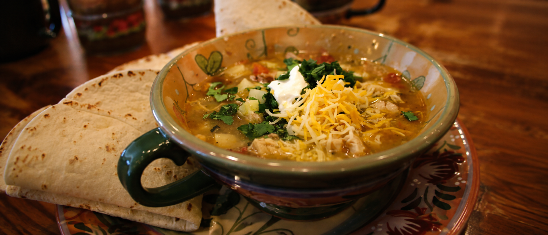 Hearty homemade green chile chicken soup topped with melted cheese, sour cream, and fresh cilantro served with warm tortillas in a rustic Southwestern restaurant setting.