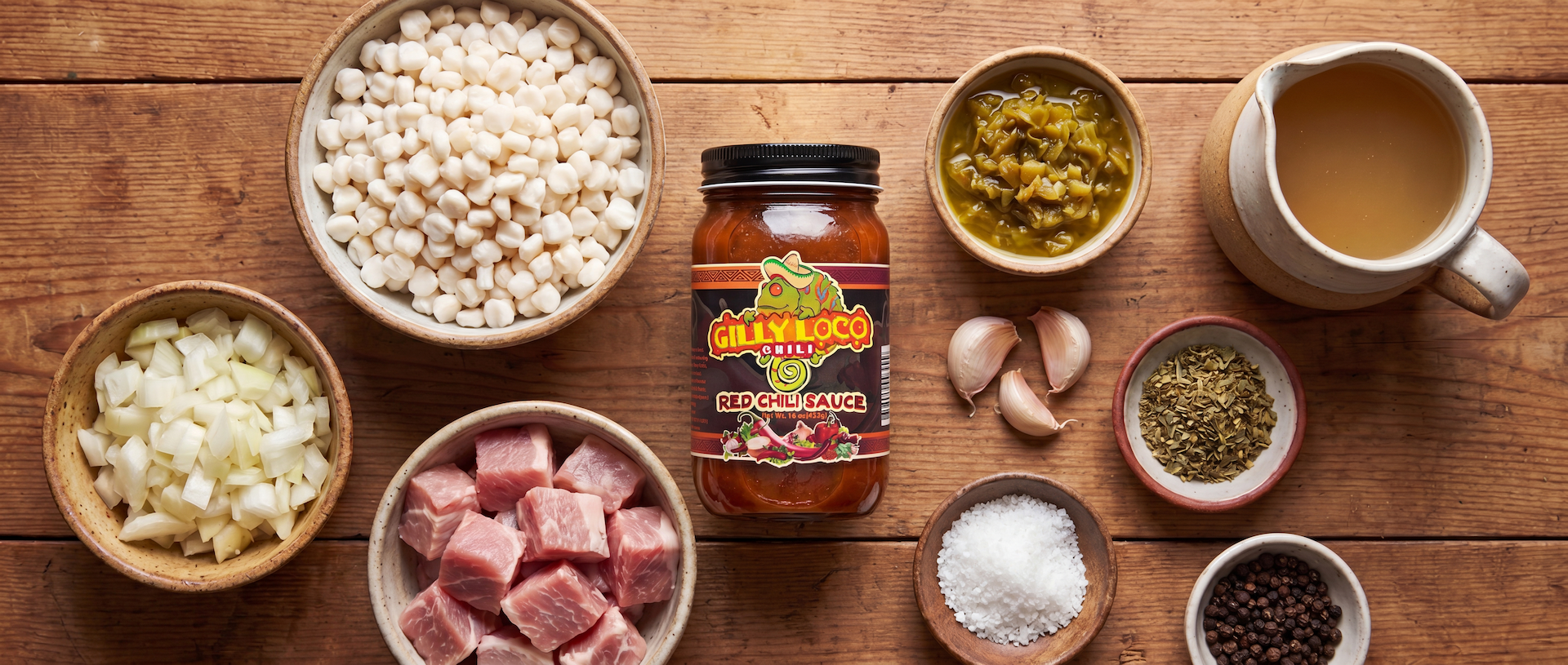 Ingredients for New Mexico Red Chile Posole including hominy, pork, onion, garlic, green chile, oregano, chicken stock, and Gilly Loco Red Chile Sauce arranged on a rustic wooden table.