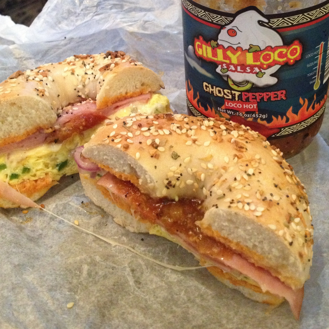 Photo of a bagel sandwich cut in half to reveal layers of cream cheese, sliced tomatoes, onions, and crisp lettuce, topped with a dollop of Gilly Loco salsa. The salsa adds a vibrant red color and a hint of spice, enhancing the fresh ingredients sandwiched between the toasted bagel halves.