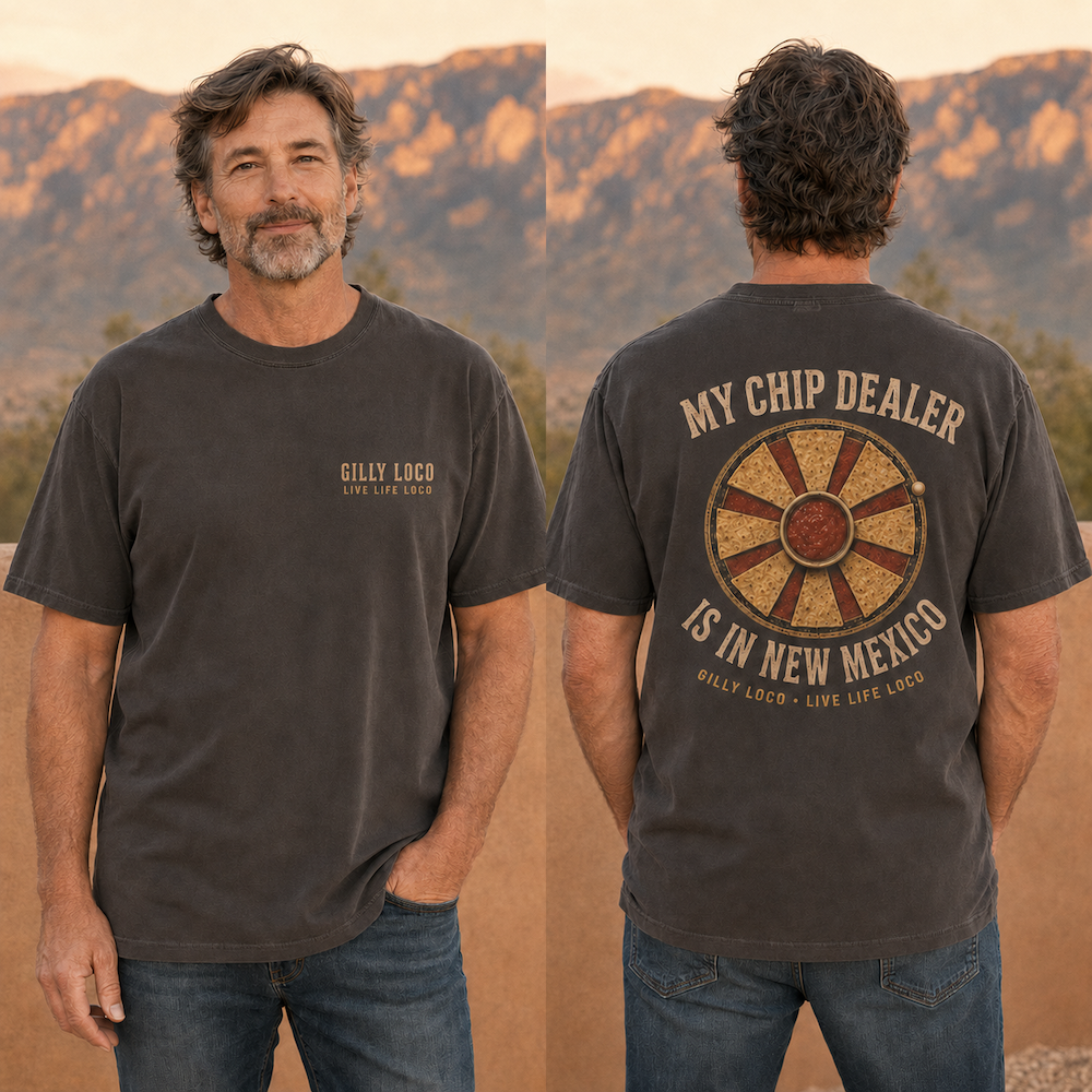 A man is shown outdoors in front of a desert mountain landscape, modeling a dark charcoal gray graphic t-shirt. The front view shows a small 'GILLY LOGO' logo on the left chest, while the back view shows a large circular wheel graphic with the text 'MY CHIP DEALER IS IN NEW MEXICO' and 'GILLY LOGO - LIVE LIFE LOGO'.