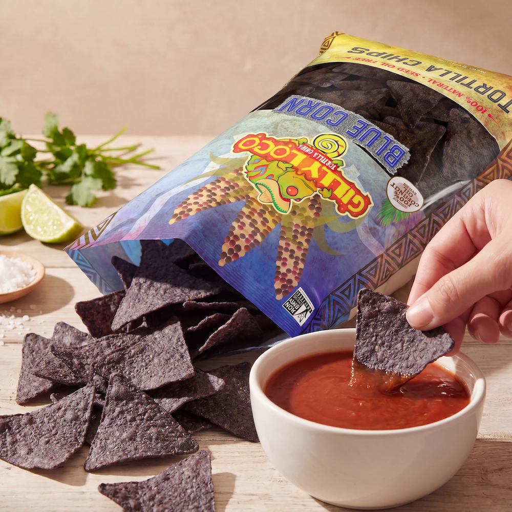 Hand dipping a Gilly Loco Blue Corn tortilla chip into a bowl of Traditional Salsa beside an open bag of Gilly Loco Blue Corn tortilla chips on a rustic tabletop with lime and cilantro