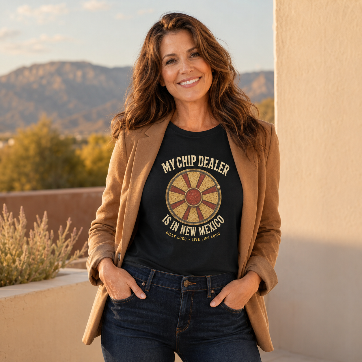 A woman wearing a black graphic t-shirt with a brown blazer and dark blue jeans, standing outdoors against a beige wall with mountains in the background. The t-shirt features a circular wheel graphic and the text 'MY CHIP DEALER IS IN NEW MEXICO'.
