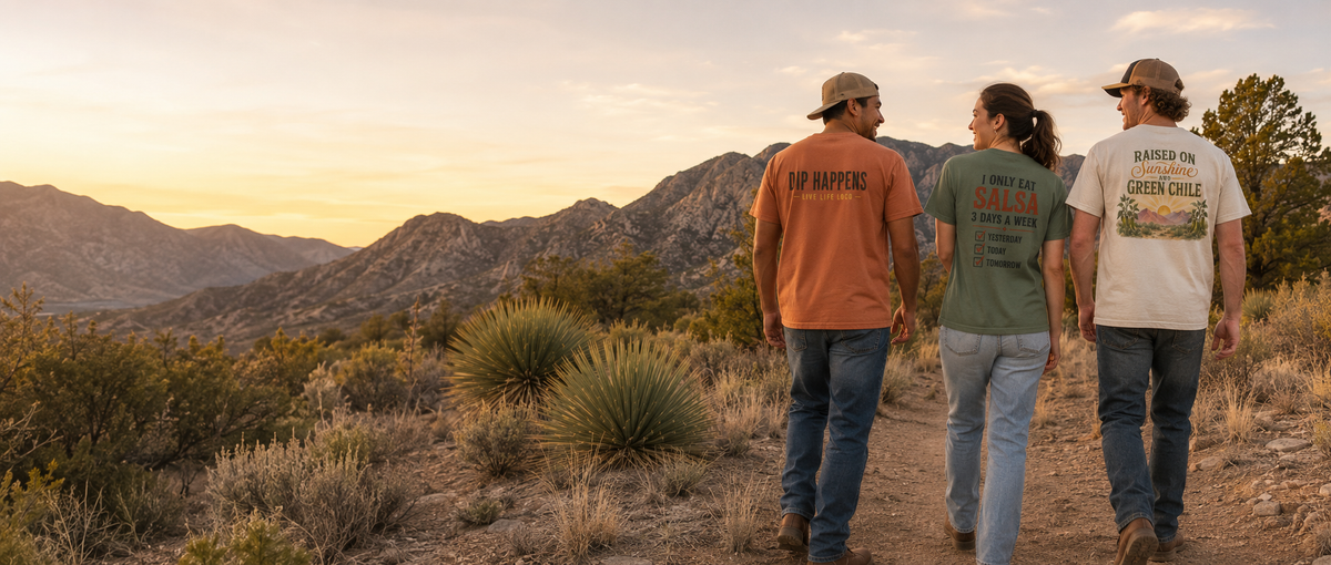 Three people walking hand in hand on a dirt trail in a desert mountain landscape at sunset, wearing graphic t-shirts. The person on the left wears an orange t-shirt with 'IF HAPPEN' printed on the back, the middle person wears a green t-shirt with 'I LOVE GREEN CHILI' printed on the back, and the person on the right wears a white t-shirt with 'BARTED BY GREEN CHILI' printed on the back.